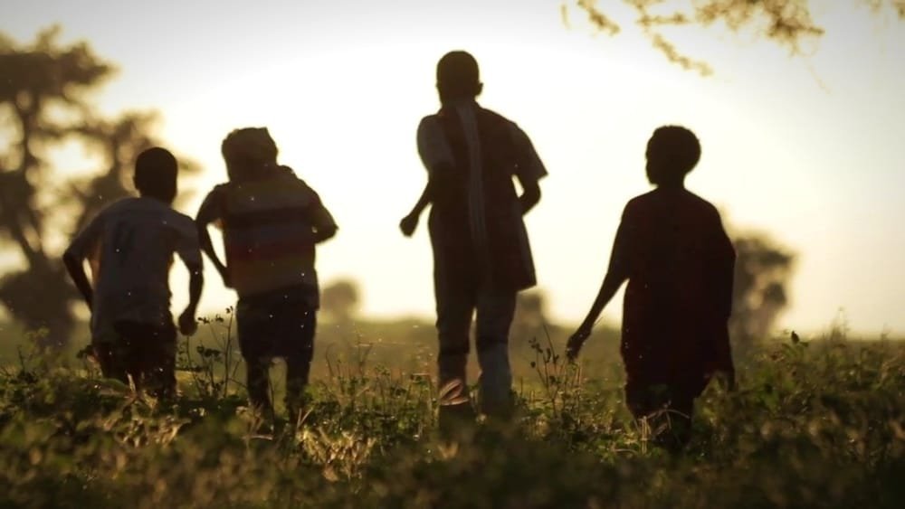Four children run across a field towards the evening sun, seen as silhouettes. A frame from Darfur in War.
