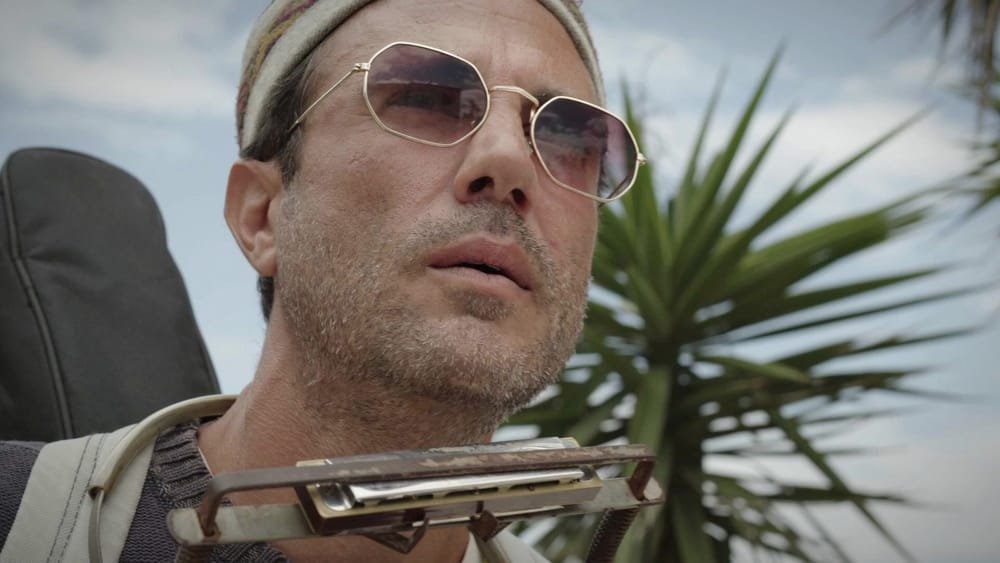 Federico, in sunglasses and a woven cap, looks up, a harmonica at his mouth. A frame from Federico the Singer.