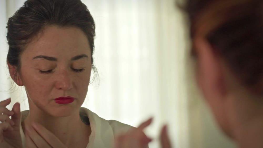 A young woman with red lipstick puts on an earring, her eyes closed. A frame from My Way.