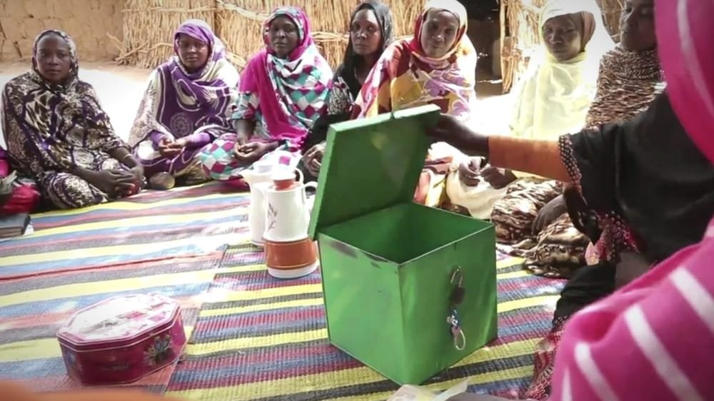 Women sit in a circle on colourful mats around an open green box. A frame from the promo for Oxfam Sudan: Advocacy Program.