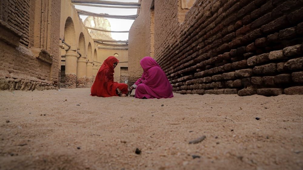 Two women in red and pink tobes sit facing each other on the sandy ground of a narrow lane between weathered brick walls. The image on the poster of Recoiling Memory of Home.
