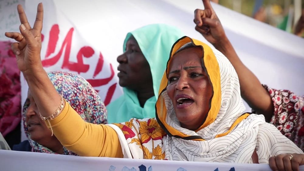 A woman in a yellow scarf raises a victory sign at a protest. A frame from Women of Sudan.
