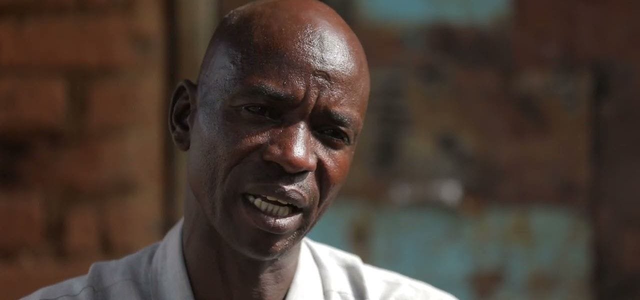 A man speaking to camera in a doorway, a weathered painted wall behind him. A frame from Darfur in War.
