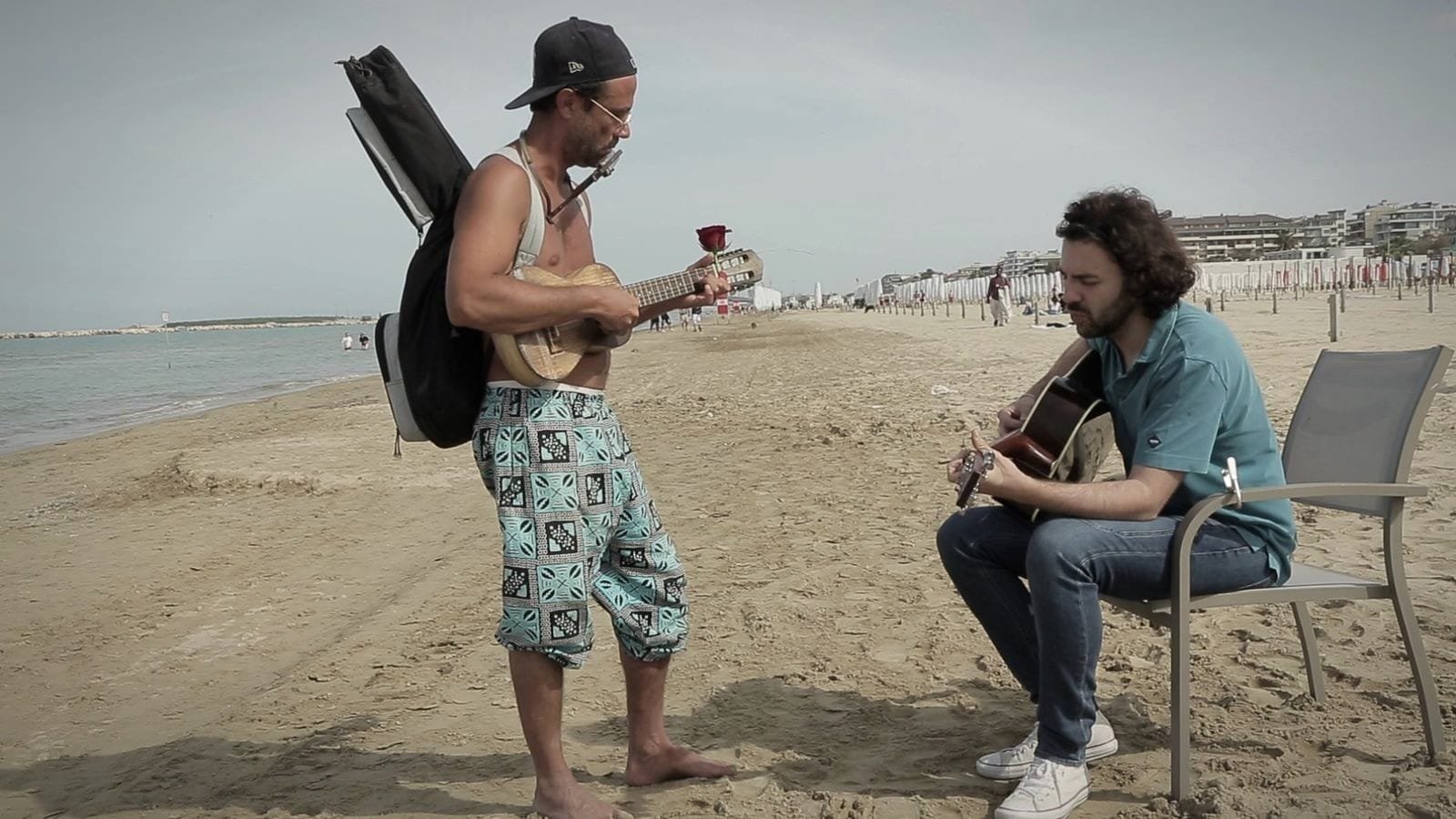 Two musicians play on the beach, one standing with a guitar case on his back, one seated. A frame from Federico the Singer.