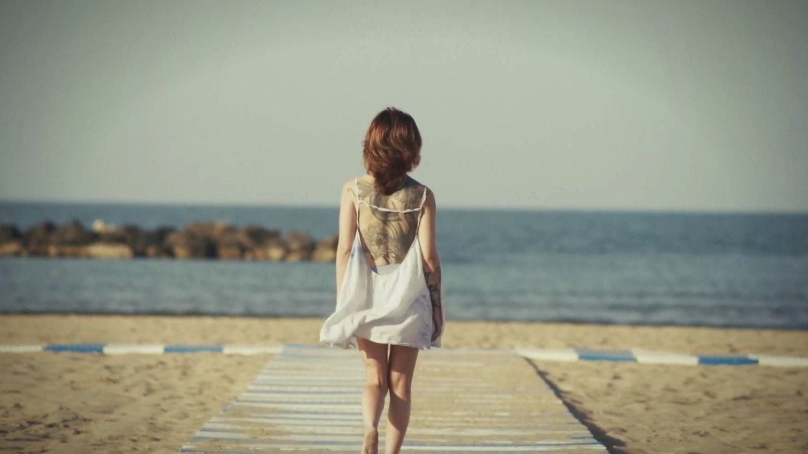 A young woman in a white dress walks across the sand towards the sea. A frame from My Way.