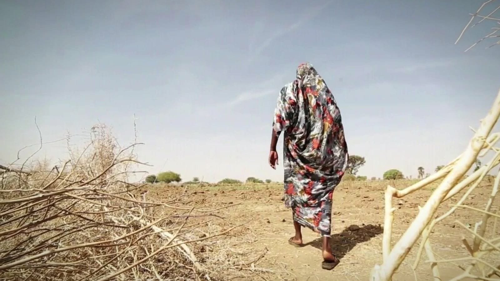 A woman in a patterned red and grey wrap walks away across dry, open land. A frame from the promo for Oxfam Sudan: Advocacy Program.