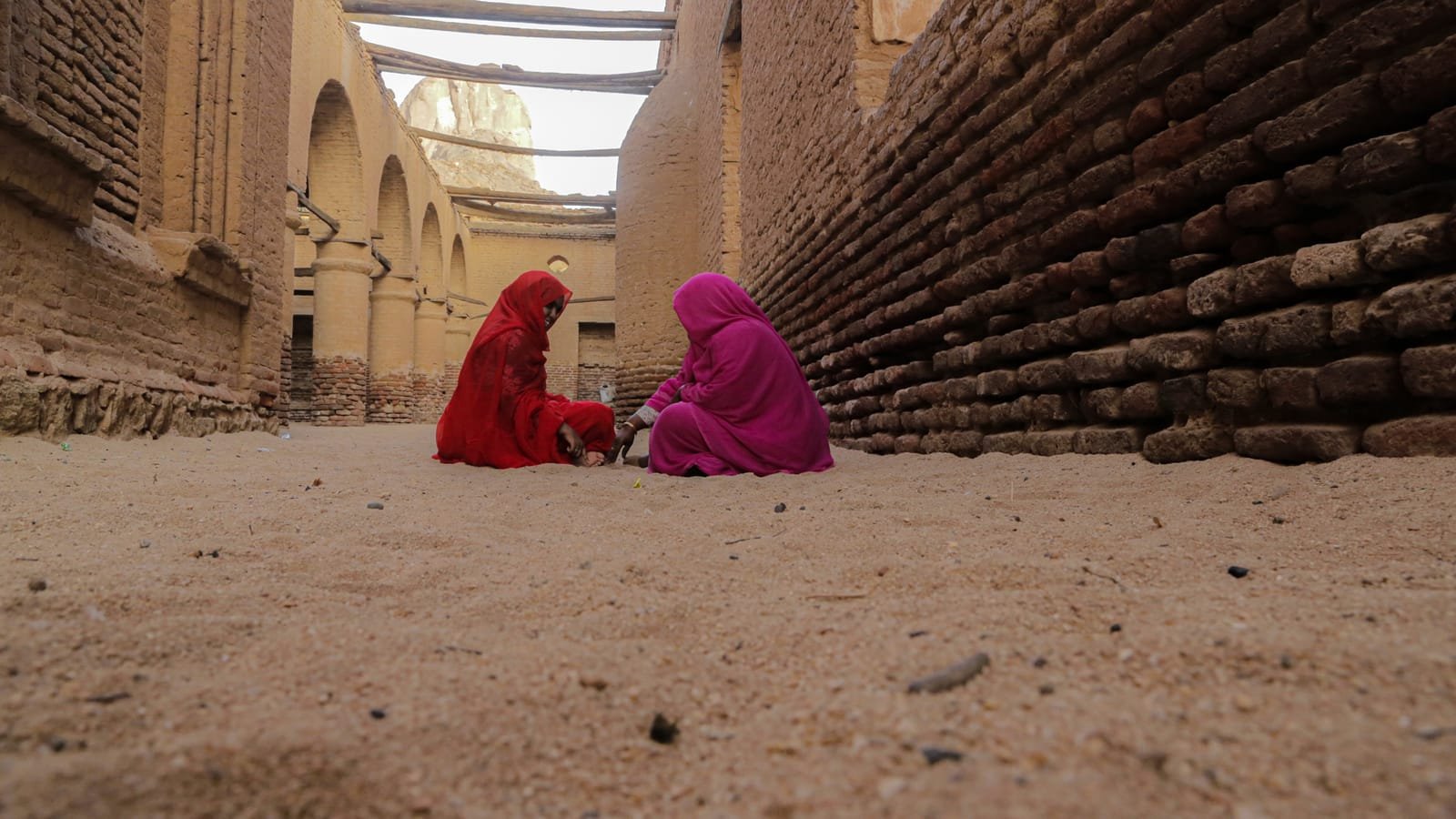 Two women in red and pink tobes sit facing each other on the sandy ground of a narrow lane between weathered brick walls. The image on the poster of Recoiling Memory of Home.