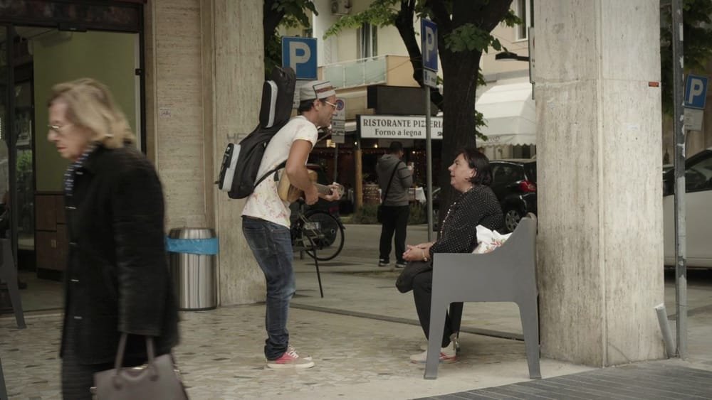 Federico plays in the street by a café while people pass by. A frame from Federico the Singer.