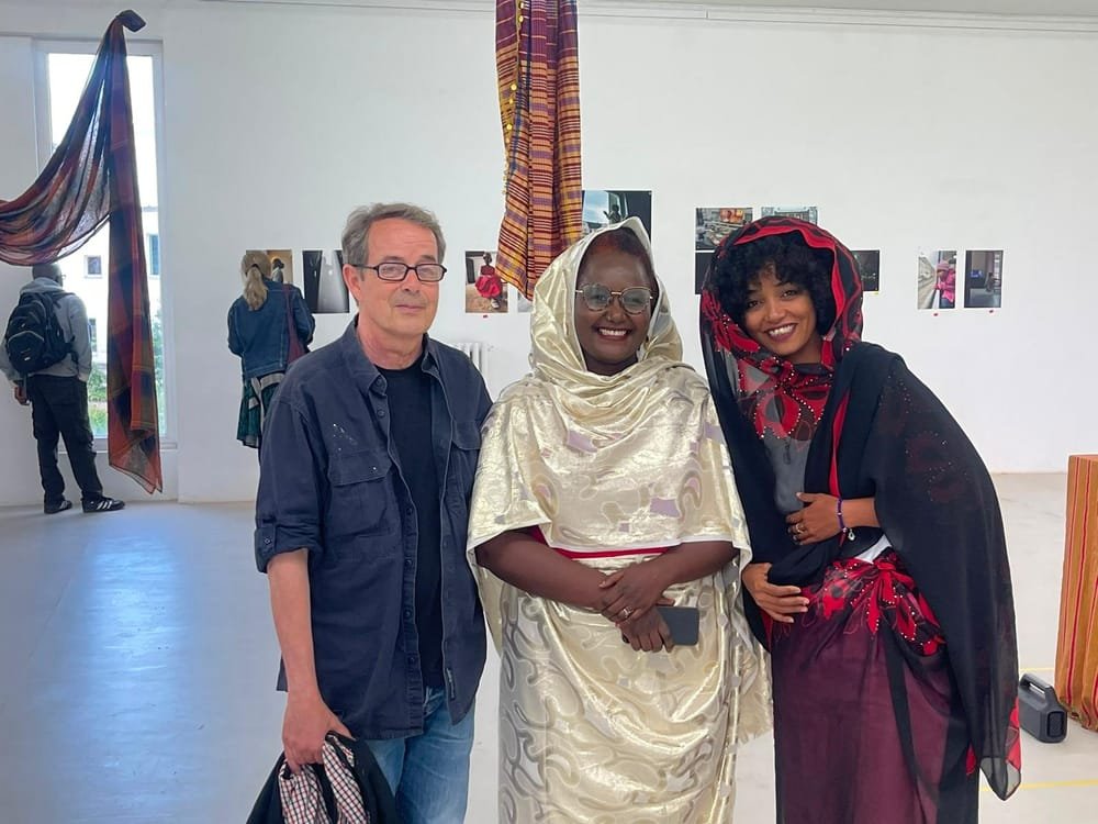 Three people pose together in the exhibition room, in front of hanging striped cloths and photographs.