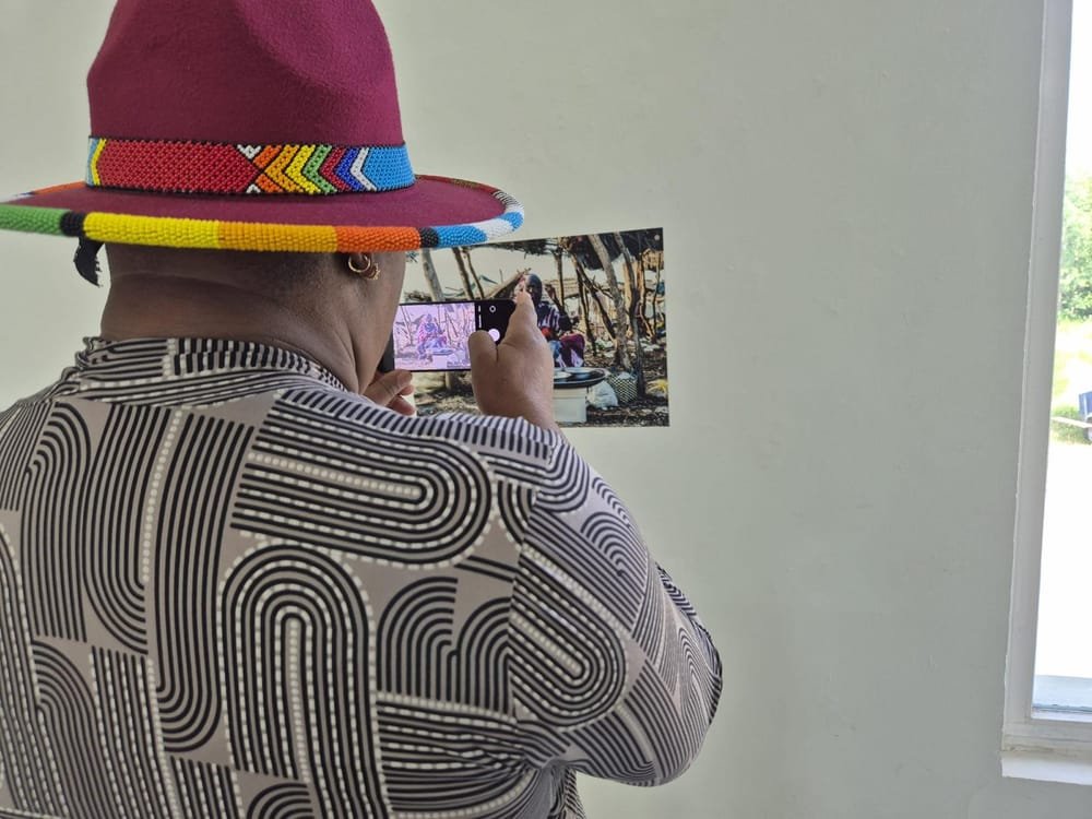 A visitor in a wide-brimmed hat with a beaded band photographs one of the pictures on the wall with a phone.