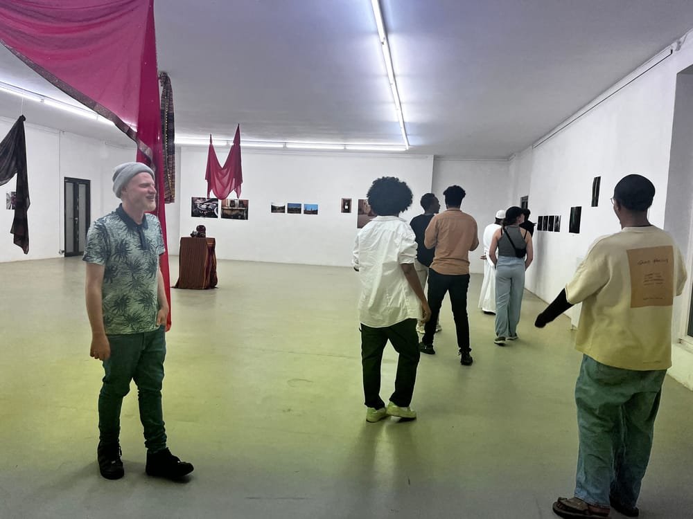 The exhibition room at Künstlerhaus Sootbörn: visitors walk past photographs on the white walls, under long red cloths hanging from the ceiling.
