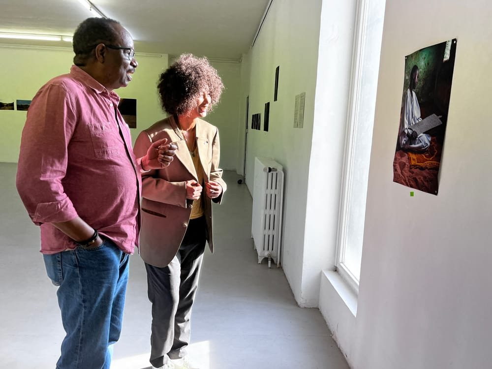 A man in a pink shirt and a woman in a beige jacket talk in front of a large photograph on the gallery wall.