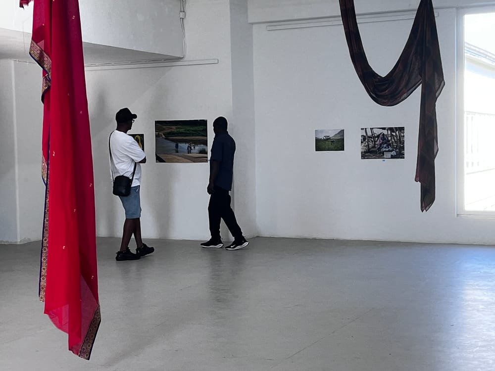 Two visitors look at photographs on a white wall, between a hanging red scarf and a long dark cloth.
