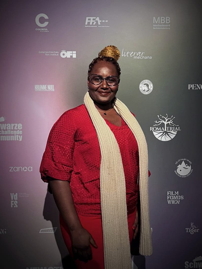Salma Abusamra in a red top and a long cream scarf, smiling in front of a wall of sponsor logos.