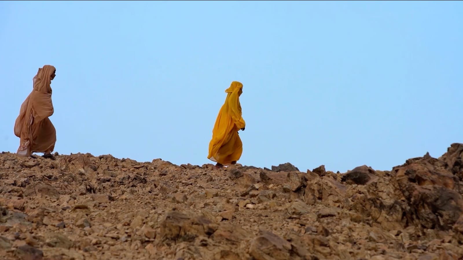 Two Sudanese women in ochre and marigold tobes walking along a rocky ridge under an open sky. A frame from the documentary Survivors.