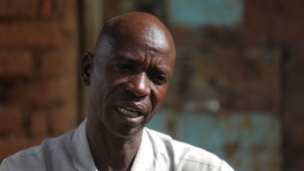 A man speaking to camera in a doorway, a weathered painted wall behind him. A frame from Darfur in War.