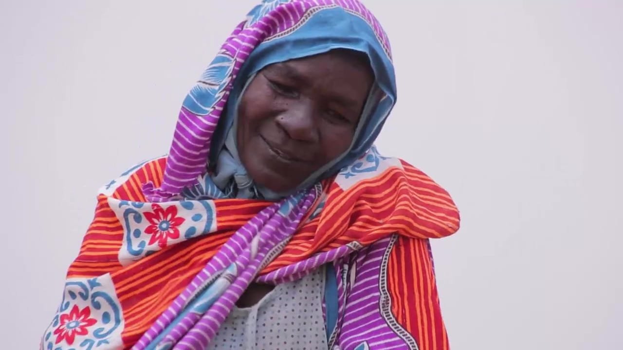 A woman in a striped purple and orange wrap looks down and smiles. A frame from the promo for El Fasher and North Darfur.