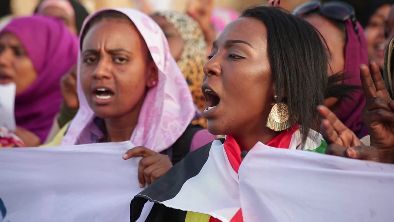 Two women chant at a protest in Sudan, holding a banner and a Sudanese flag. A frame from Women of Sudan.